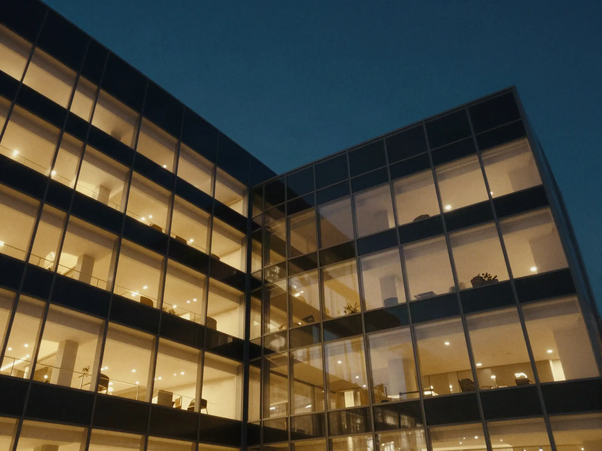 Edifício comercial de vidro visto de baixo ao anoitecer, com as janelas iluminadas.